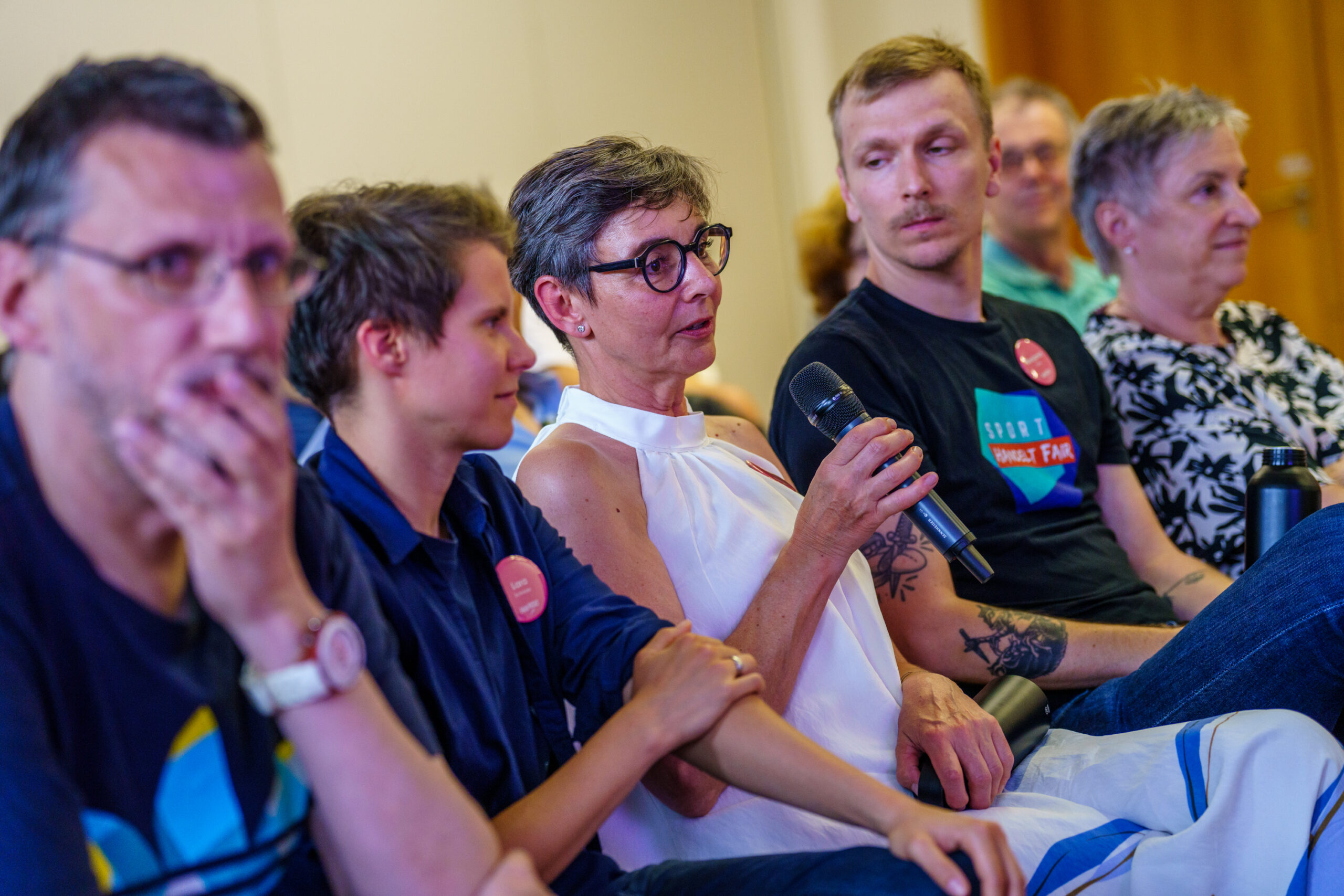 Fünf Menschen sitzen nebeneinander in einem Raum, die mittlere Person hält ein Mikrofon in der Hand und spricht. Im Hintergrund sitzen weitere Menschen.