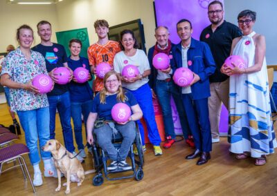 Eine Gruppe Menschen steht in einem Raum, eine Person im Rollstuhl und mit Hund an der Leine befindet sich vor der Gruppe. Alle halten lilafarbene Fußbälle mit weißen Smilies in der Hand und lächeln fröhlich in die Kamera.