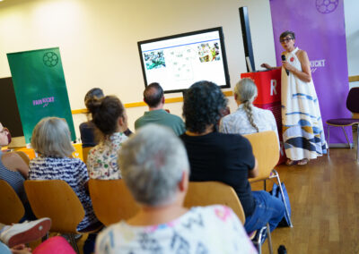 Eine Frau mit Brille in langem Kleid steht an einem Stehtisch vor einer Gruppe sitzender Menschen. Im Hintergrund stehen grüne und lila Roll-ups mit der Aufschrift Fair Kickt.