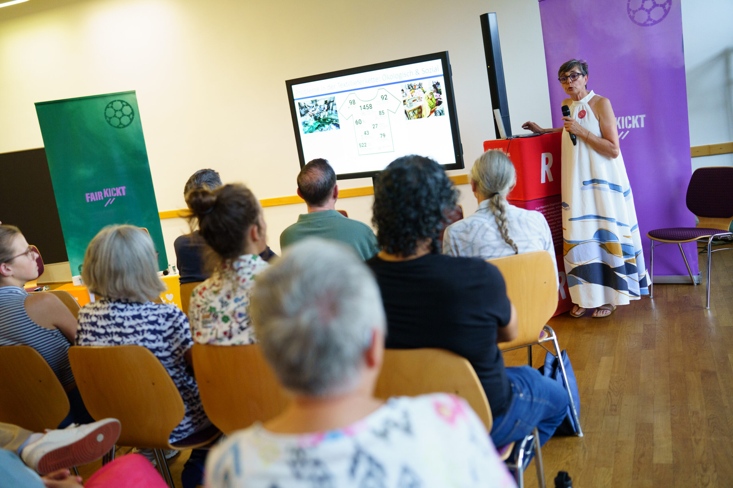 Eine Frau mit Brille in langem Kleid steht an einem Stehtisch vor einer Gruppe sitzender Menschen. Im Hintergrund stehen grüne und lila Roll-ups mit der Aufschrift Fair Kickt.