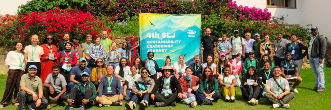Gruppenbild mit Menschen aus allen Teilen der Erde auf einer Wiese vor üppig blühenden Büschen. In der Mitte ist ein Plakat sichtbar mit der Aufschrift 4. Sustainable Leadership Journey.